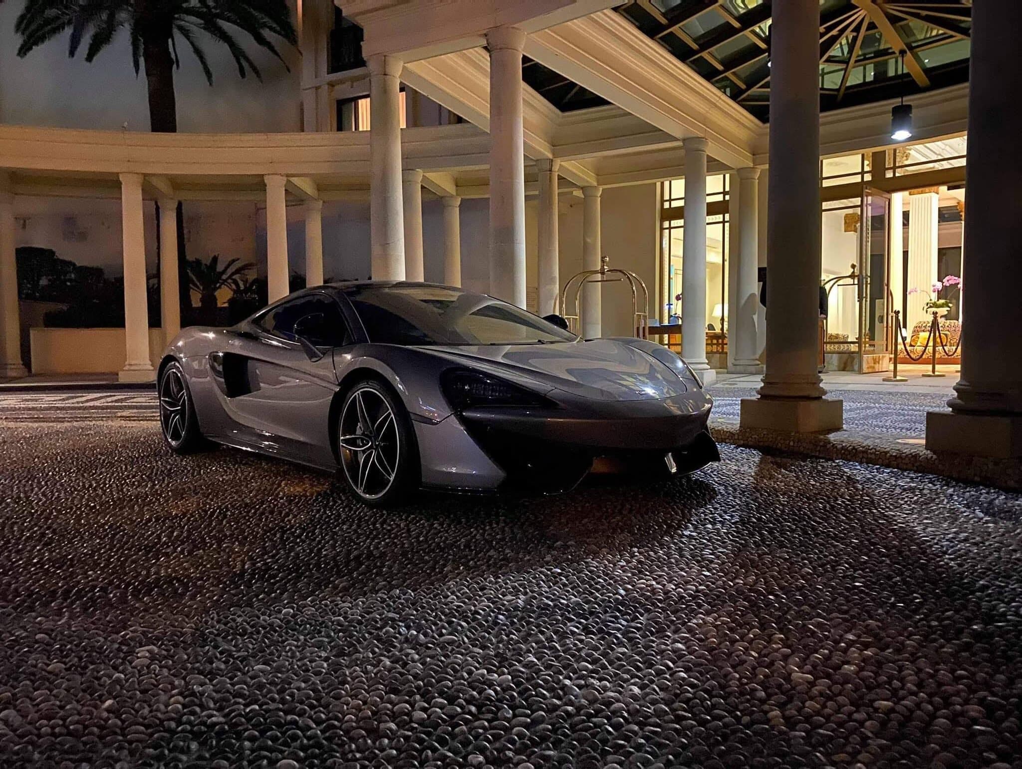 A grey McLaren 570S parked on a pebbled forecourt beneath a colonnaded entrance at night